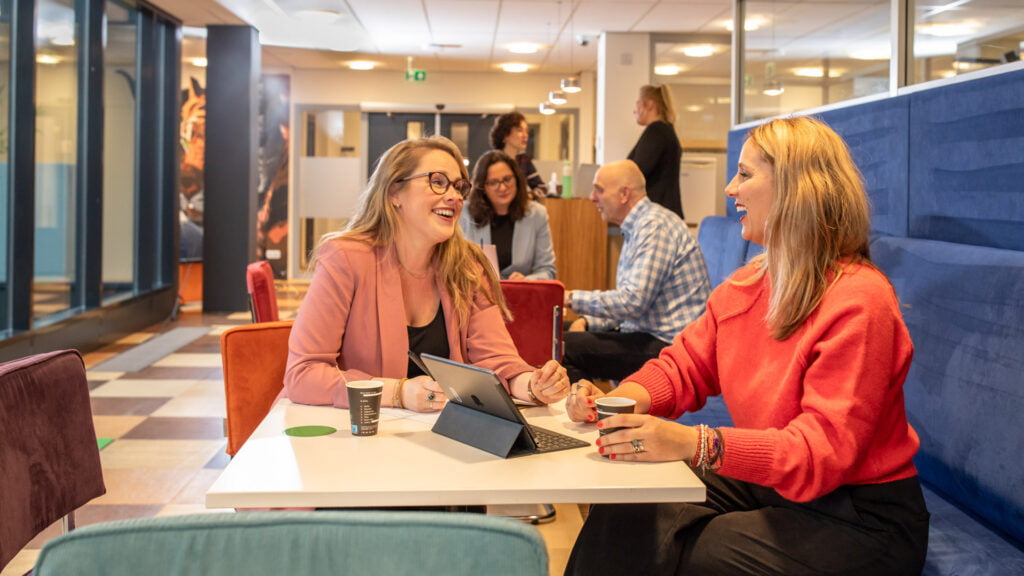 Twee medewerkers met elkaar in gesprek met in het midden een iPad en op de achtergrond andere medewerkers die met elkaar praten