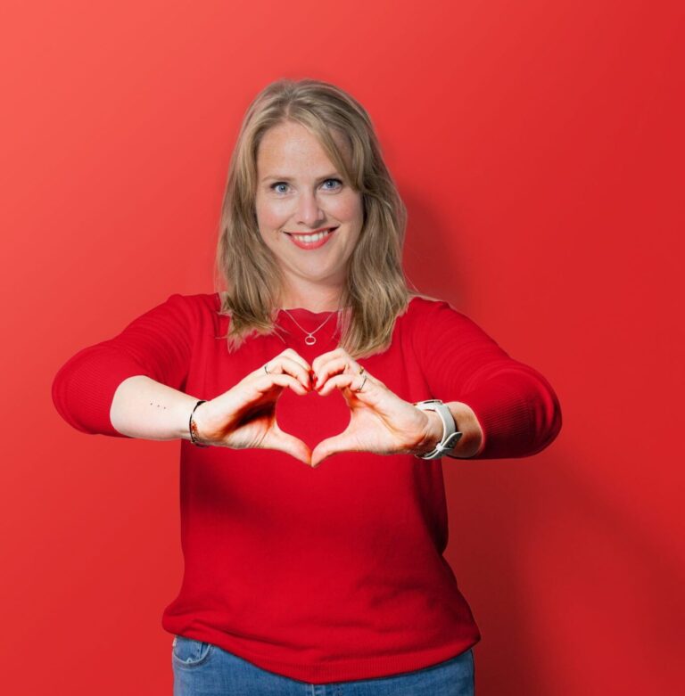 Een collega van de Sociale Dienst Drechtsteden in rode kleding en rode achtergrond die met haar handen een hartje symboliseert.