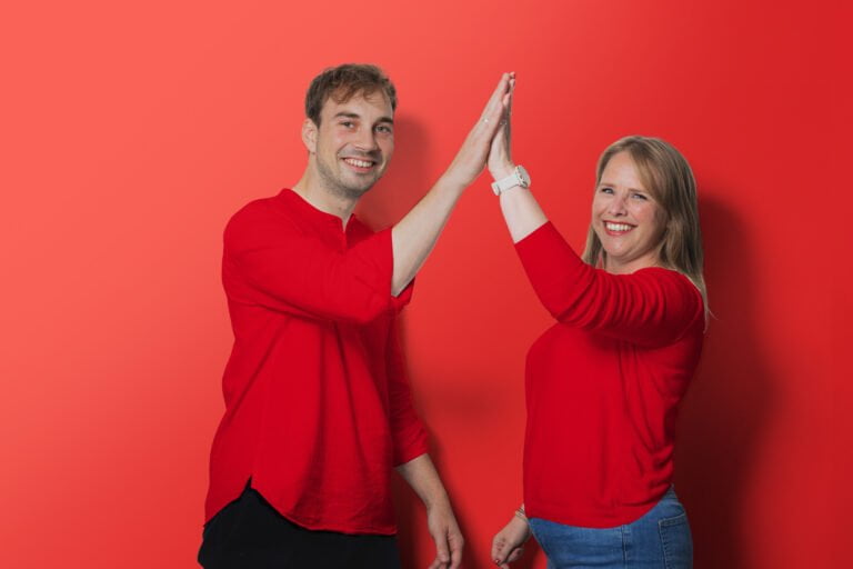 Twee collega's van de Sociale Dienst Drechtsteden in rode kleding en rode achtergrond die elkaar een high five geven.